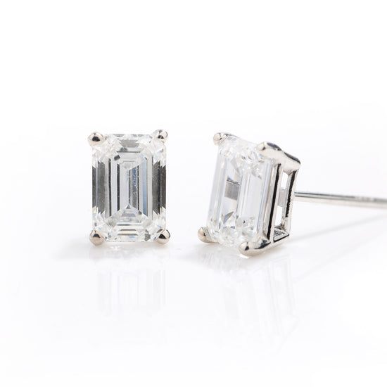A pair of emerald-cut diamond stud earrings in a silver four-prong setting, shown on a clean white reflective surface.
