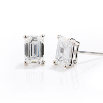 A pair of emerald-cut diamond stud earrings in a silver four-prong setting, shown on a clean white reflective surface.