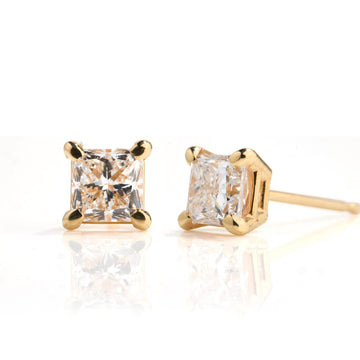 A pair of gold stud earrings with princess-cut clear gemstones, shown on a reflective white surface. One earring is facing forward, displaying the four-prong setting and the square-cut clear stone, while the other is angled to show the side profile and post.