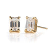 A pair of gold stud earrings featuring rectangular baguette-cut clear gemstones in four-prong settings, shown on a reflective white surface.