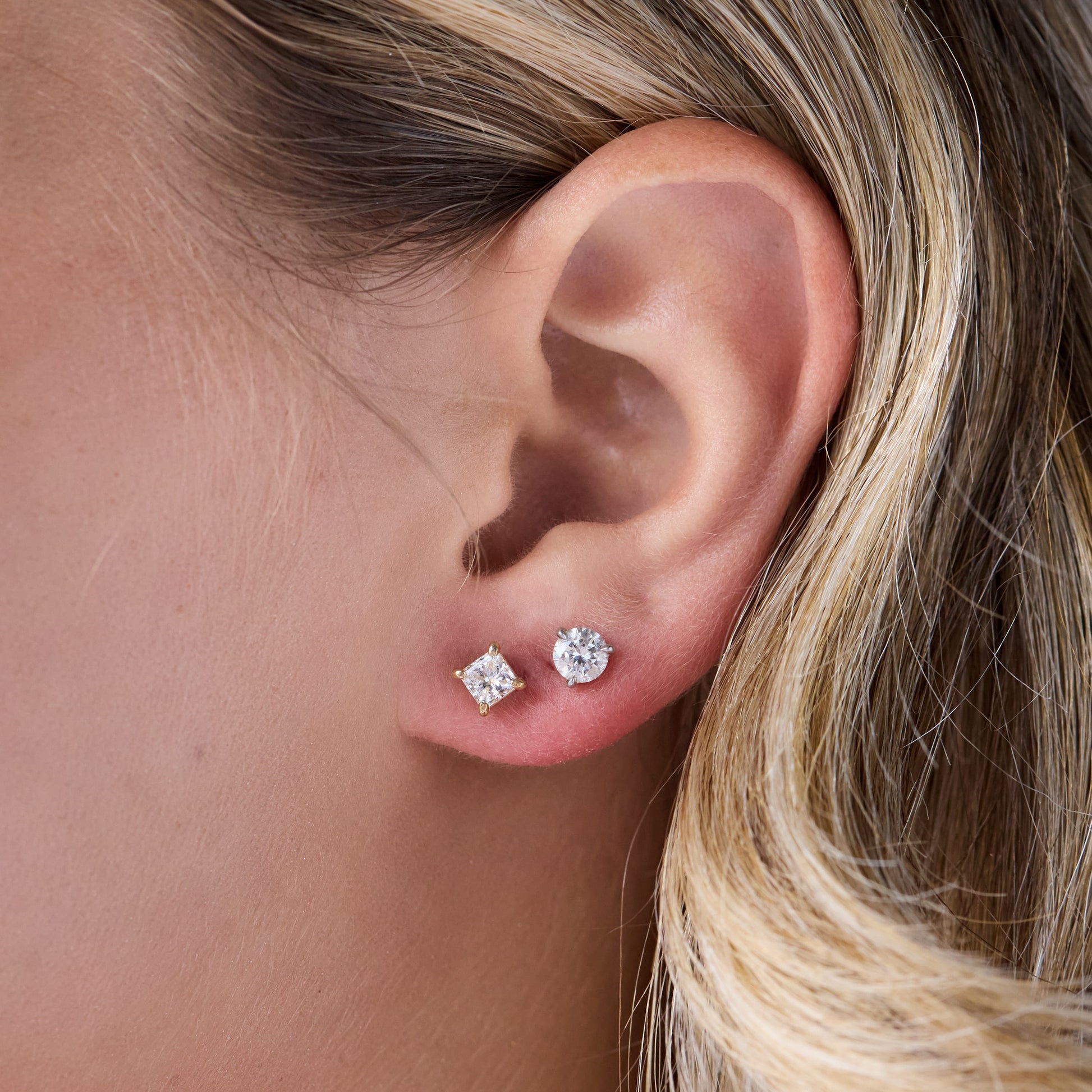 A close-up of a person's ear wearing two small stud earrings: one square-cut clear stone and one round clear stone in gold-toned settings. The person has blonde hair, and the background is mostly filled by skin and hair.