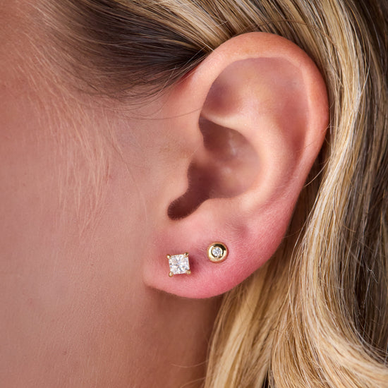Close-up of a person's ear wearing two stud earrings: a square-cut clear gemstone stud on the lobe and a small round gold stud above it.