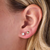 Close-up of a person's ear wearing two stud earrings: a square-cut clear gemstone stud on the lobe and a small round gold stud above it.