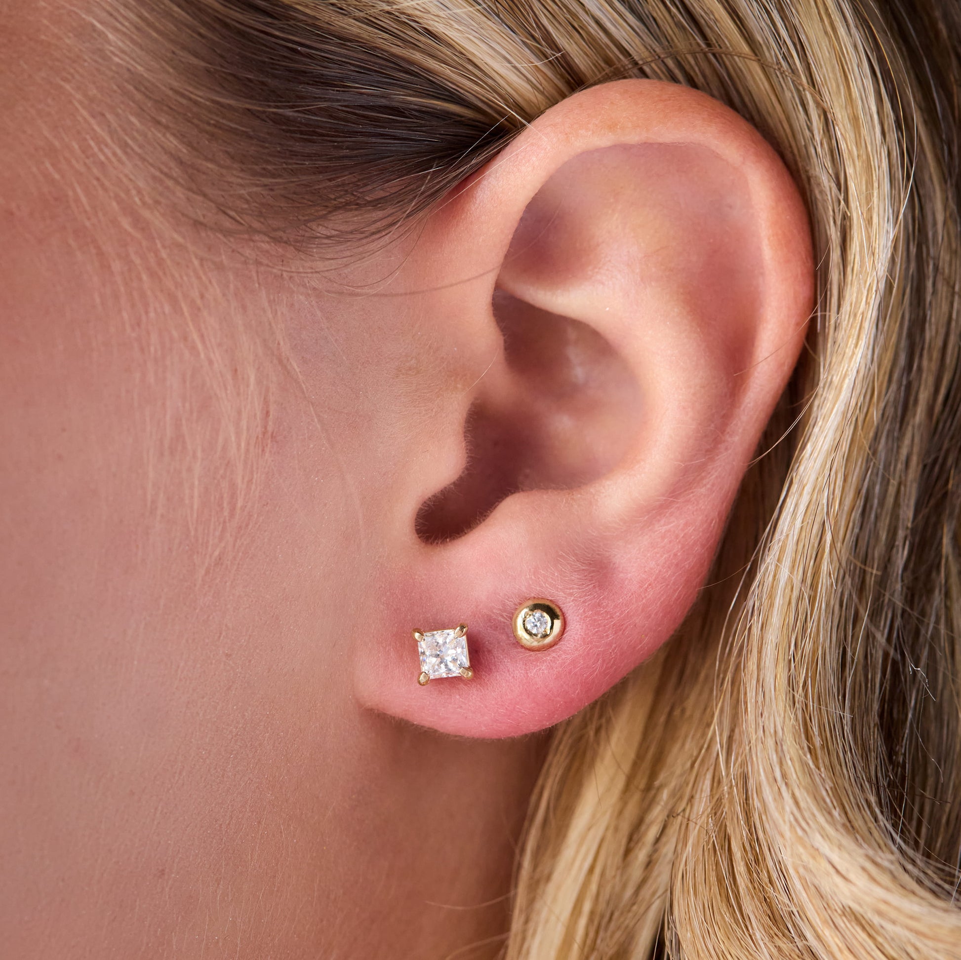 Close-up of a person's ear wearing two stud earrings: a square-cut clear gemstone stud on the lobe and a small round gold stud above it.