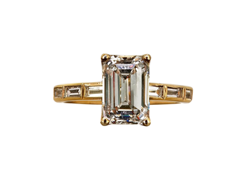 A yellow gold ring featuring a large emerald-cut clear center stone with baguette side stones, set against a plain white background.