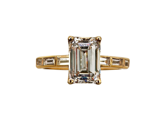 A yellow gold ring featuring a large emerald-cut clear center stone with baguette side stones, set against a plain white background.