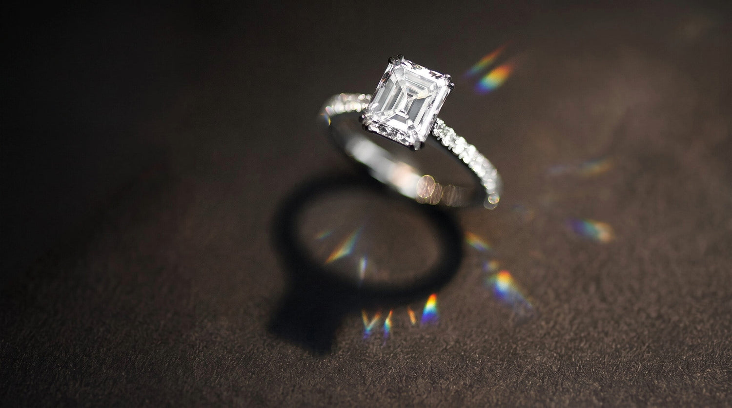 A silver engagement ring with a rectangular emerald-cut diamond center stone and a pavé band, resting on a dark textured surface with a soft shadow and rainbow light leaks.